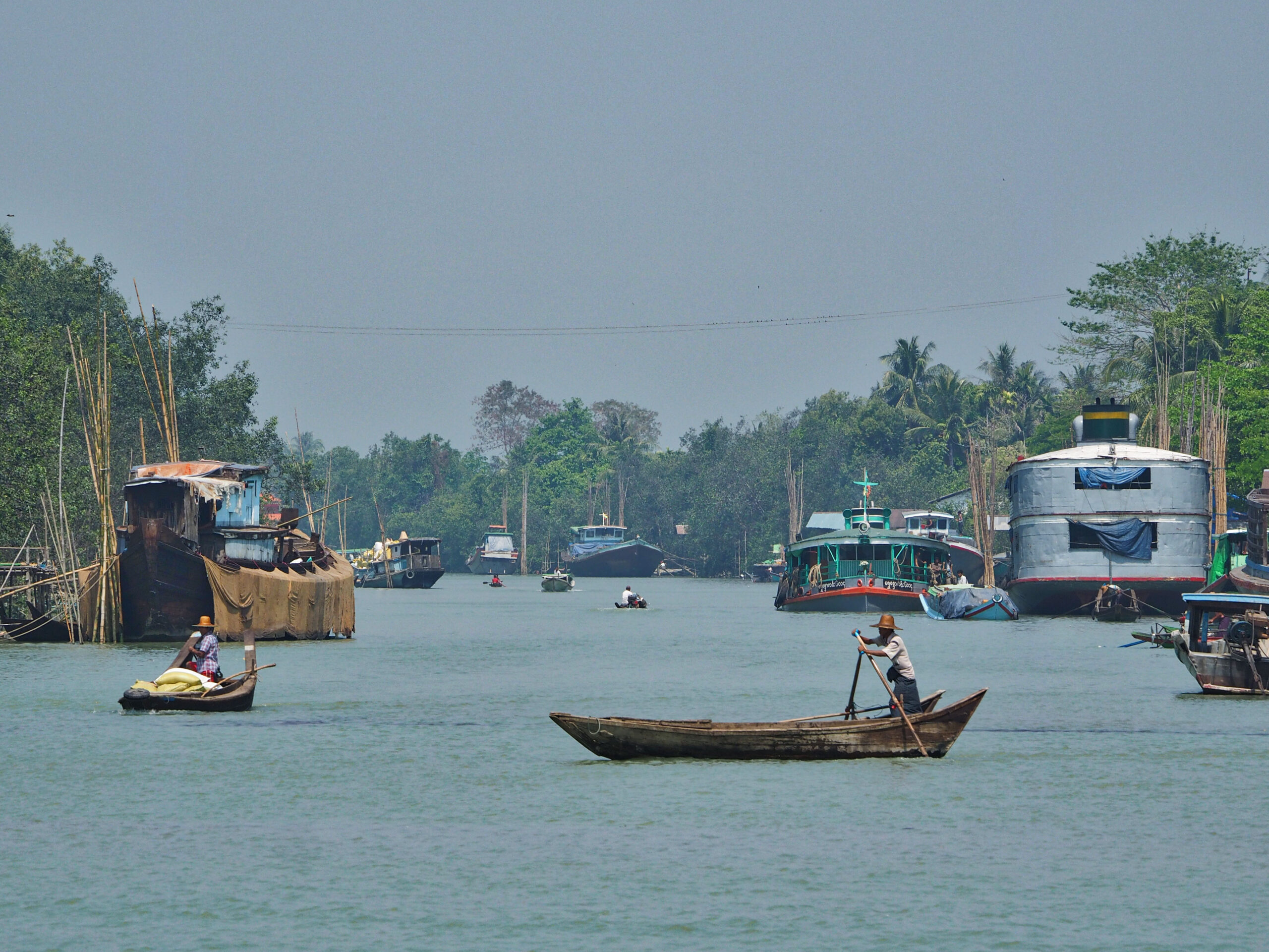 Traditionelle Holzboote und Lastenschiffe auf dem Irrawaddy-Fluss in Südostasien, Fischer mit Strohhüten rudern durch das Wasser