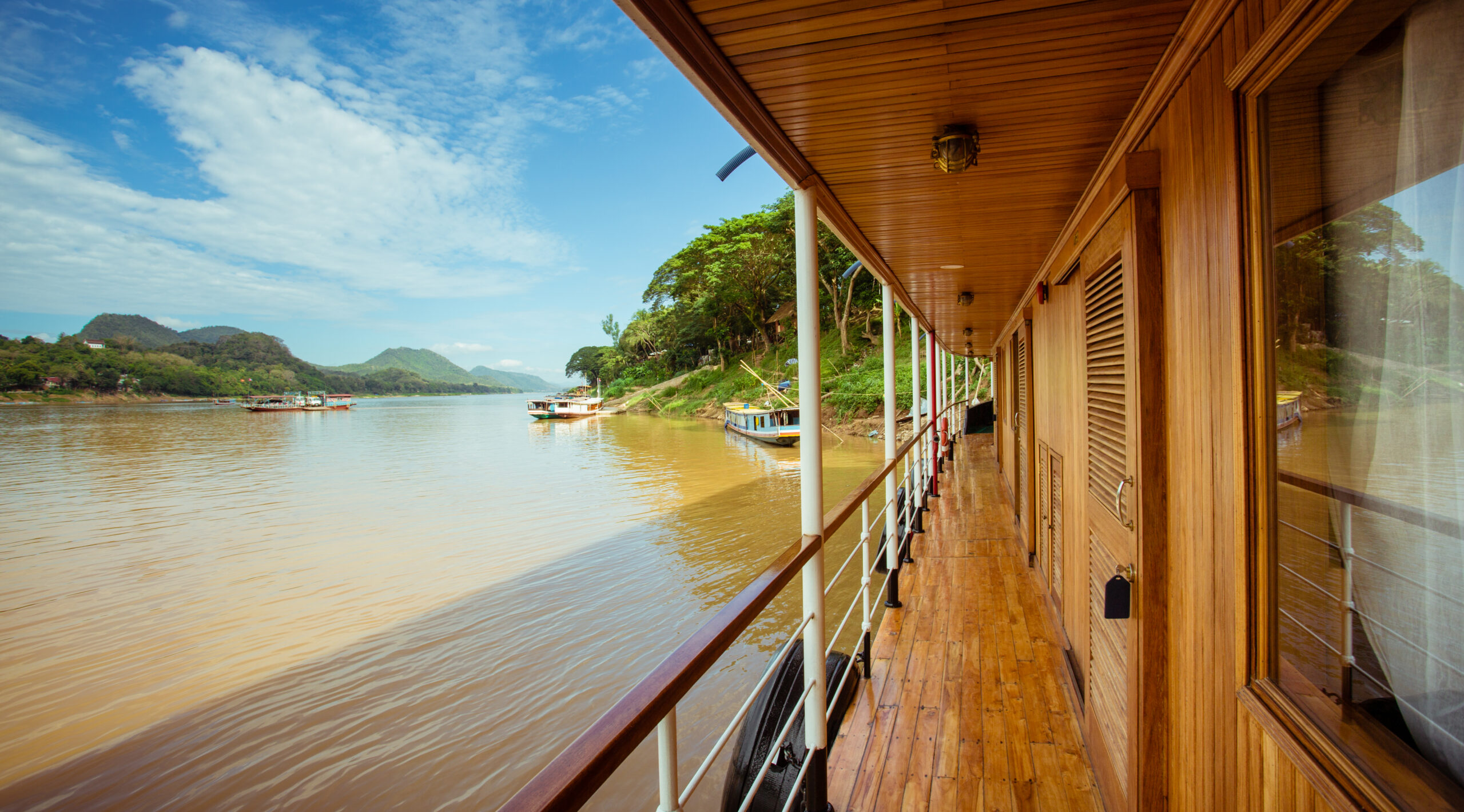 Holzterasse eines Flusskreuzfahrtschiffs mit Blick auf ruhigen Fluss Irrawaddy und grüne Berglandschaft an der Küste