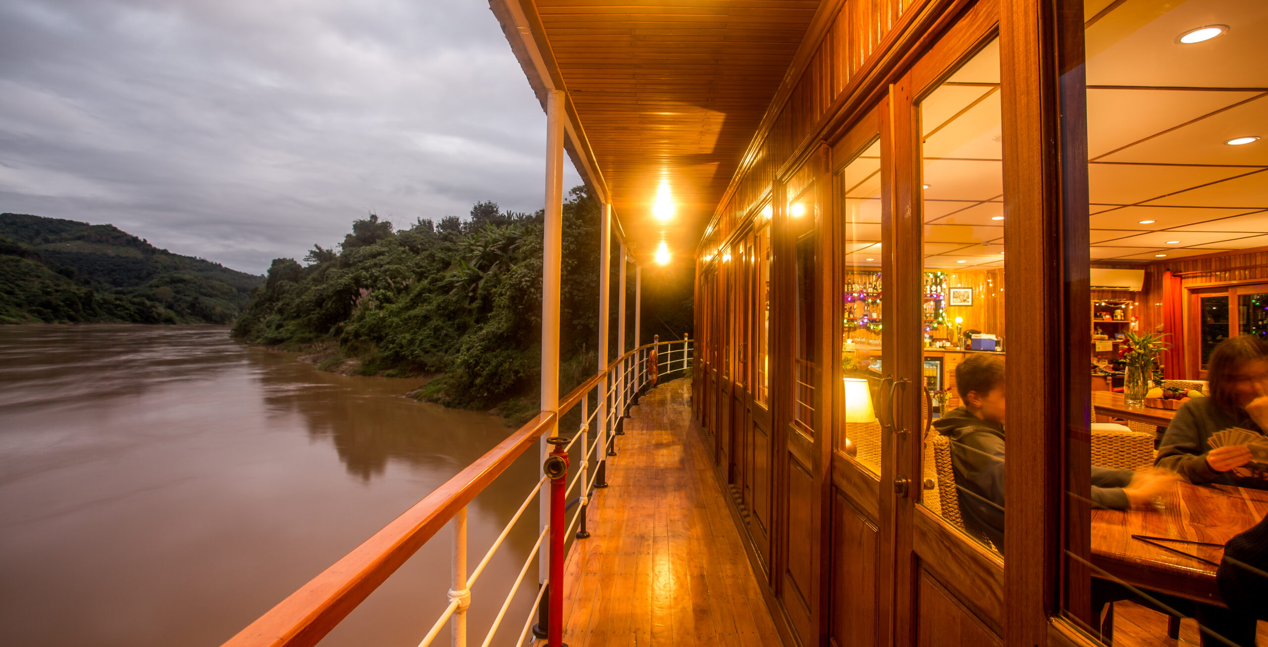 Veranda eines Flusskreuzfahrtschiffs bei Dämmerung mit Blick auf ruhigen Fluss und grüne Uferhügel, warmes Licht von innen