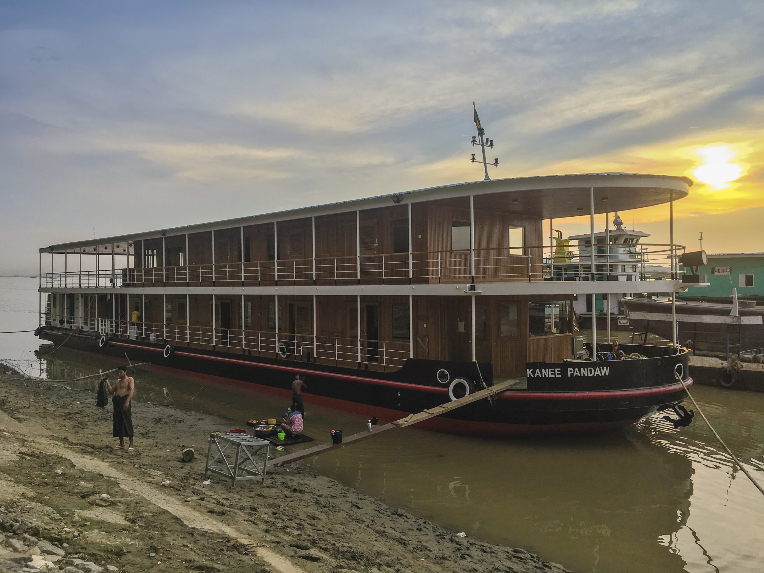 Das Flusskreuzfahrtschiff Kanee Pandaw liegt bei Sonnenuntergang am flachen Ufer, Menschen bereiten sich am Steg vor