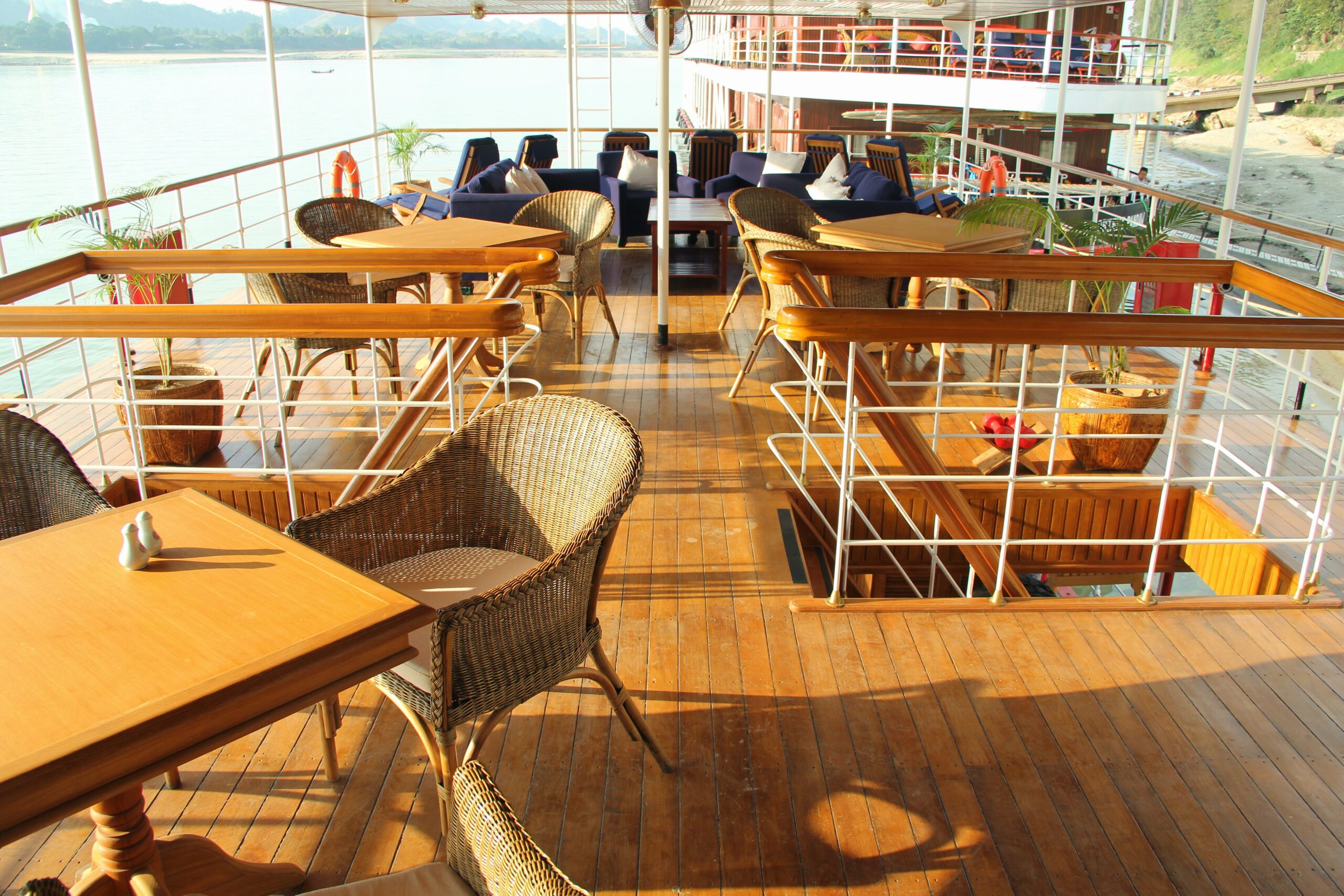 Sonnendeck des Flusskreuzfahrtschiffs RV Kna Byoo Pandaw mit Holztischen und Rattanstühlen bei warmem Sonnenlicht, Blick auf ruhigen Fluss