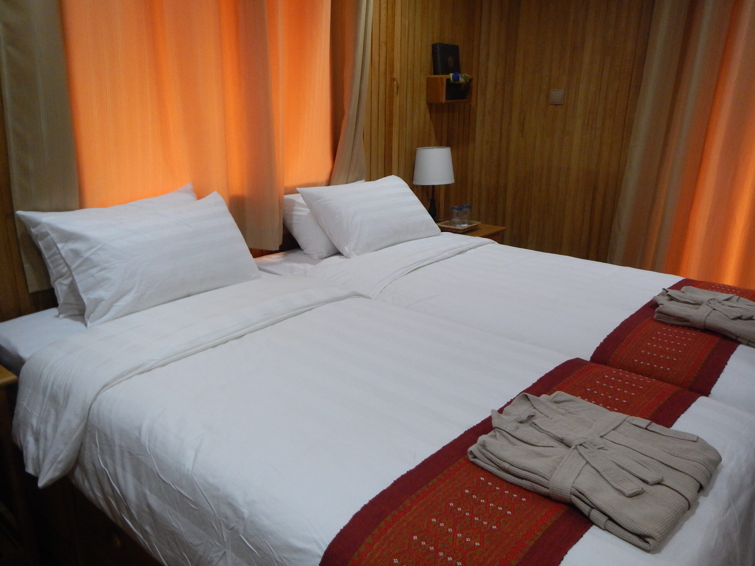 Cozy Flusskreuzfahrt Kabine mit weiß bezogenem Doppelbett, hölzernen Wänden, orangefarbenen Vorhängen und gefalteten Bademänteln auf dekorativen roten Läufern