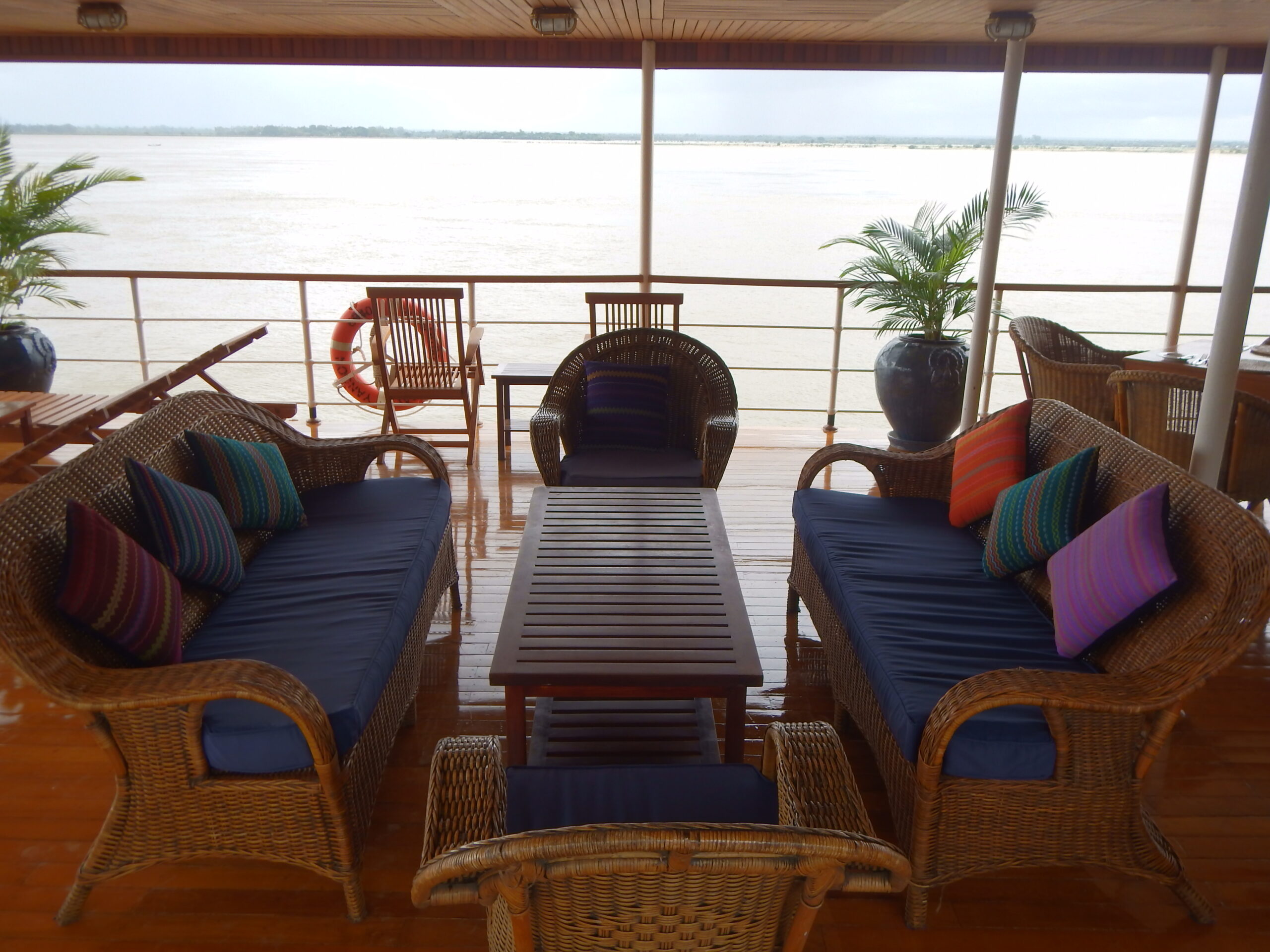 Überdachter Loungebereich auf Flusskreuzfahrtschiff mit blauen Rattansofas und bunten Kissen, Blick auf breiten Fluss