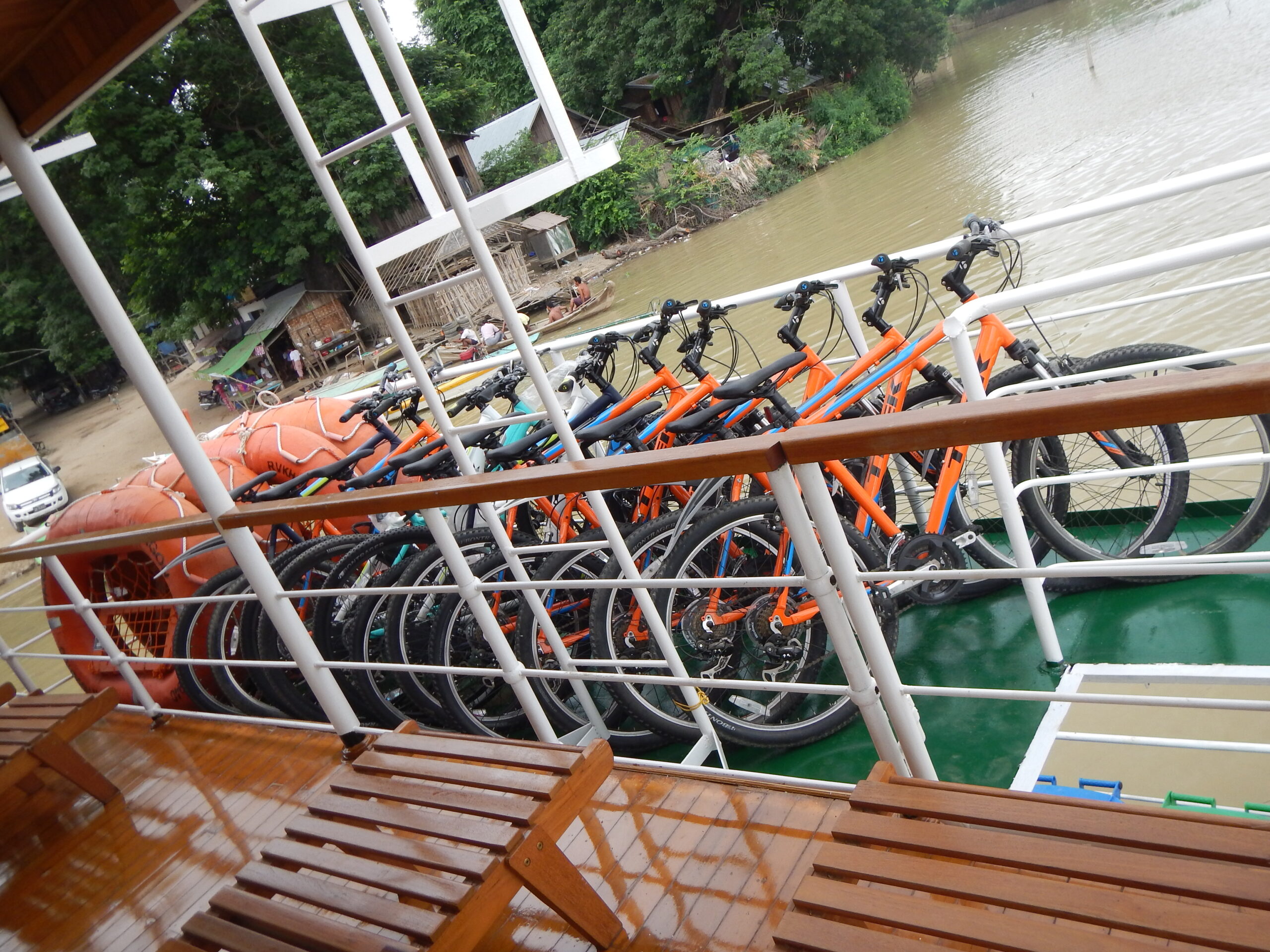 Orange Mountainbikes gereiht auf dem Deck eines Flusskreuzfahrtschiffs mit Blick auf Flussufer und Häuser im Hintergrund